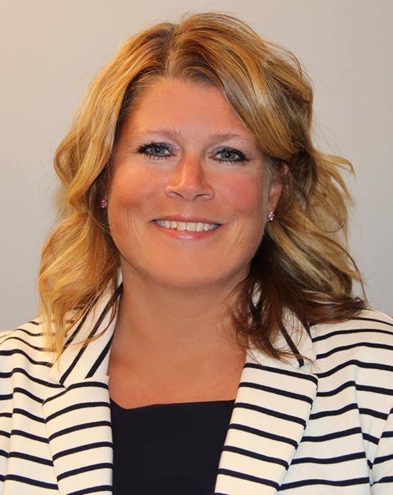 A woman with blonde hair smiles at the camera, wearing a striped blazer.