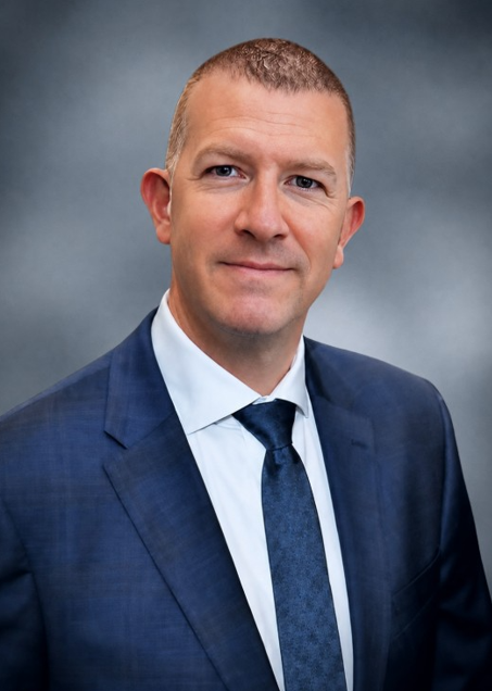A man in a blue suit and tie looks directly at the camera.