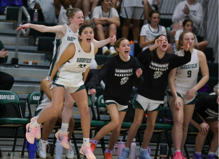 Young women in green and white basketball uniforms cheer enthusiastically from the sidelines.