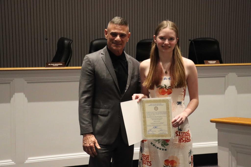 A man in a suit and a young woman in a patterned dress stand together holding a certificate.