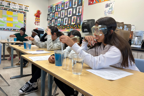 Students wearing safety goggles conduct a science experiment at their desks in a brightly lit classroom.