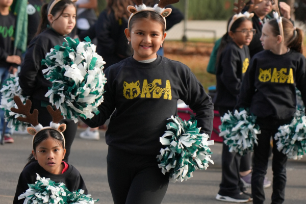 A smiling child wearing a black sweatshirt with gold lettering and holding green and white pom-poms.