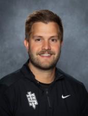 A man with a mustache smiles at the camera, wearing a black athletic jacket.