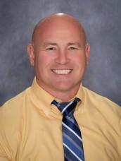 A man with a shaved head smiles at the camera, wearing a yellow shirt and tie.