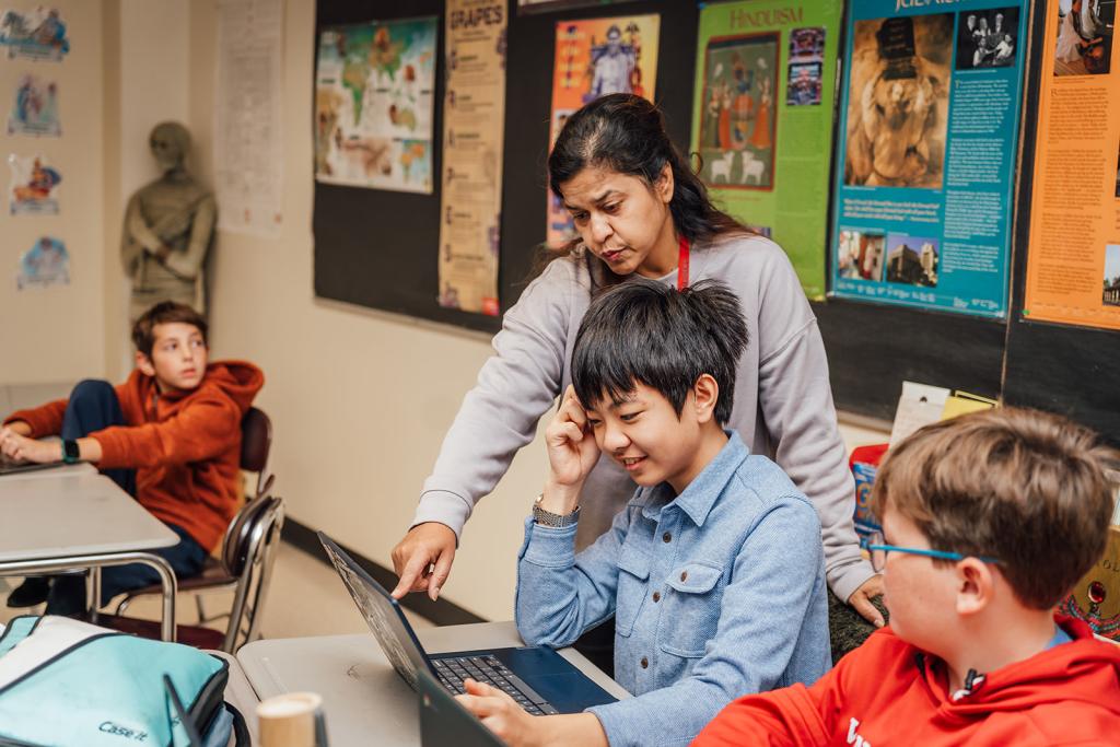 A teacher assists a student using a laptop in a classroom setting.