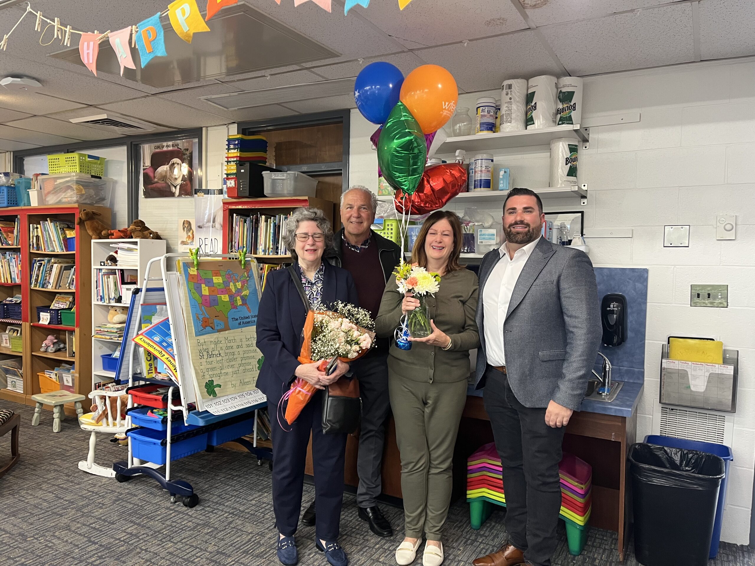 Four people stand together, holding flowers and balloons, in a library setting.
