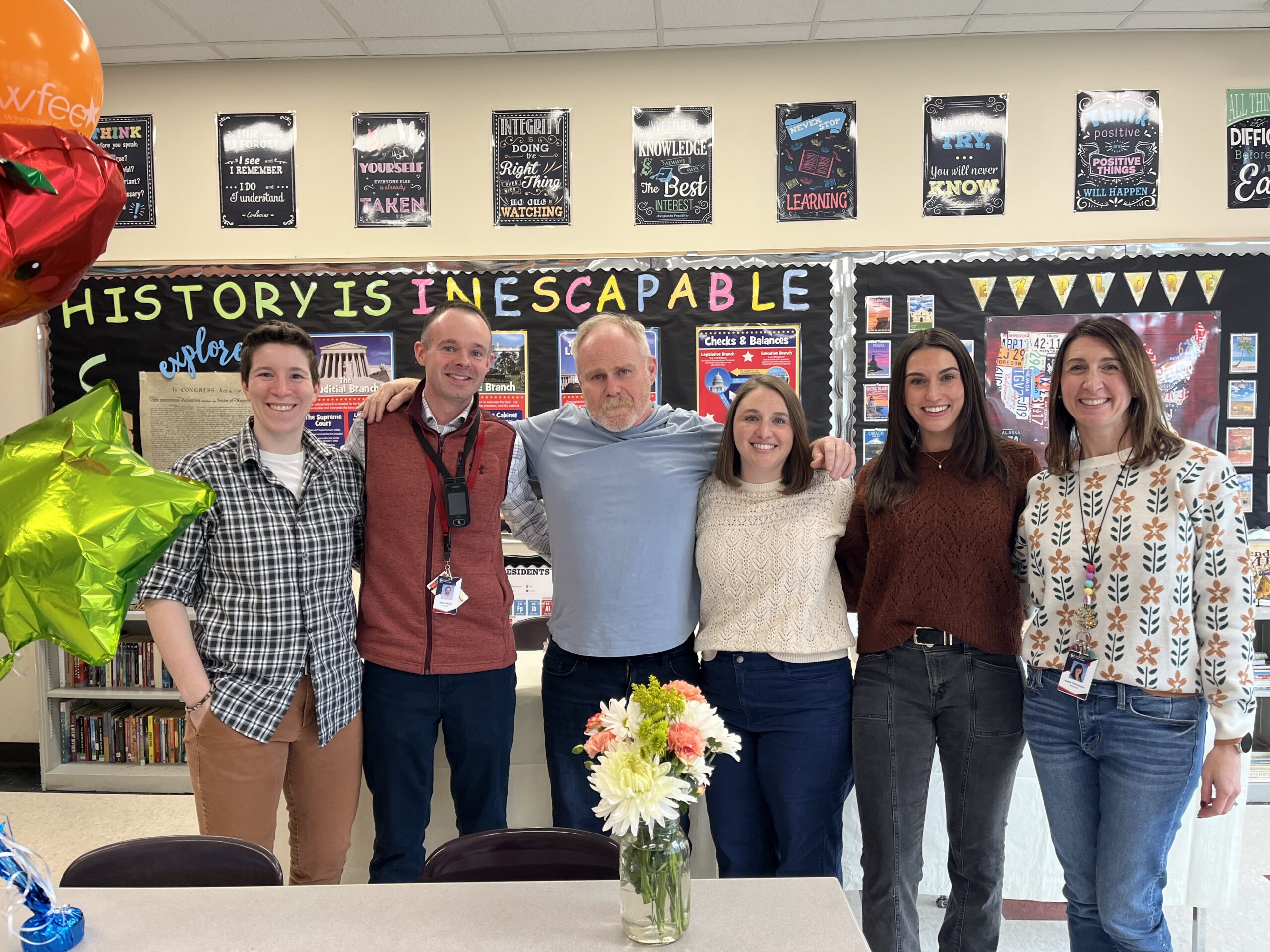 A group of six people stand together, smiling in front of a classroom backdrop.