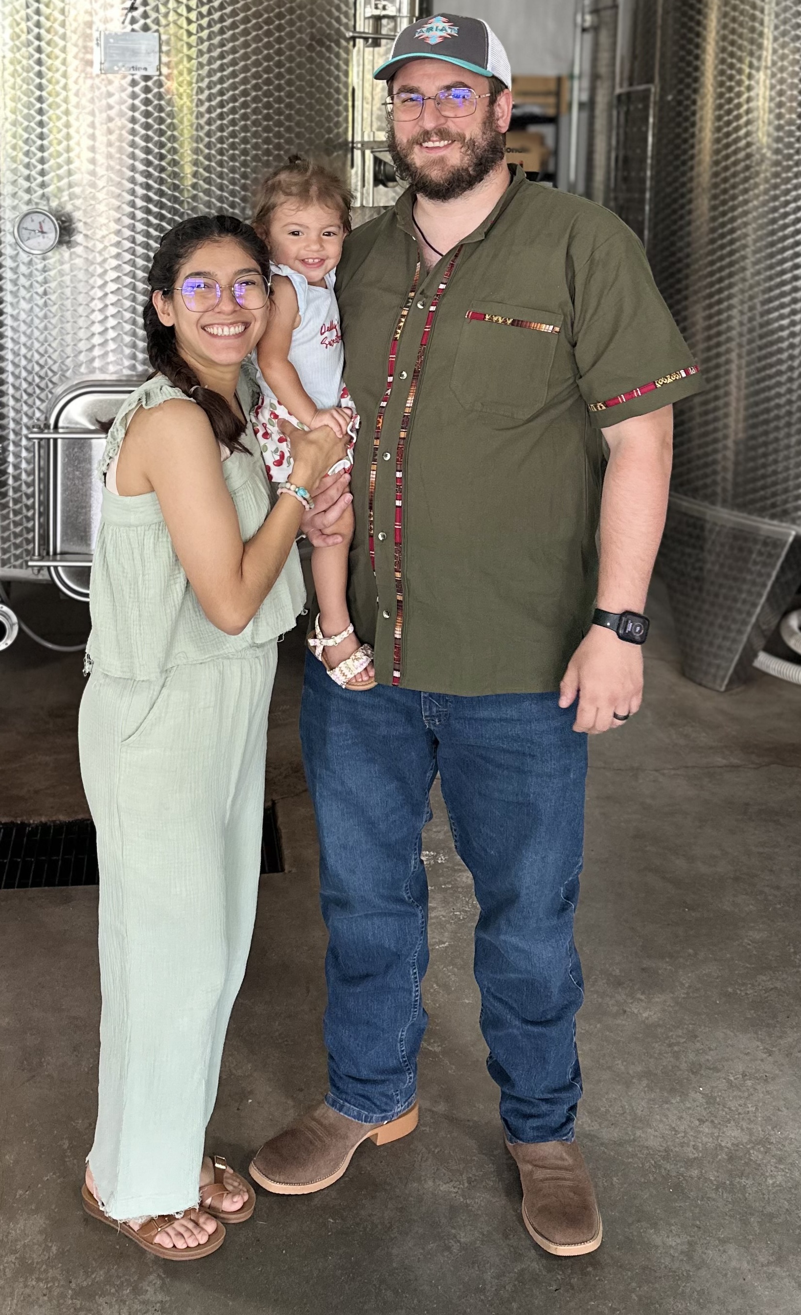 A family of three poses together inside a building with large metal tanks in the background.