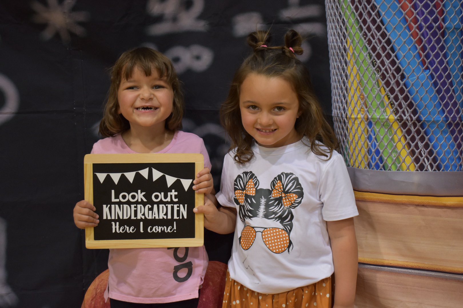 Two young girls smile, one holding a sign that reads 'Look Out Kindergarten! Here I come!'