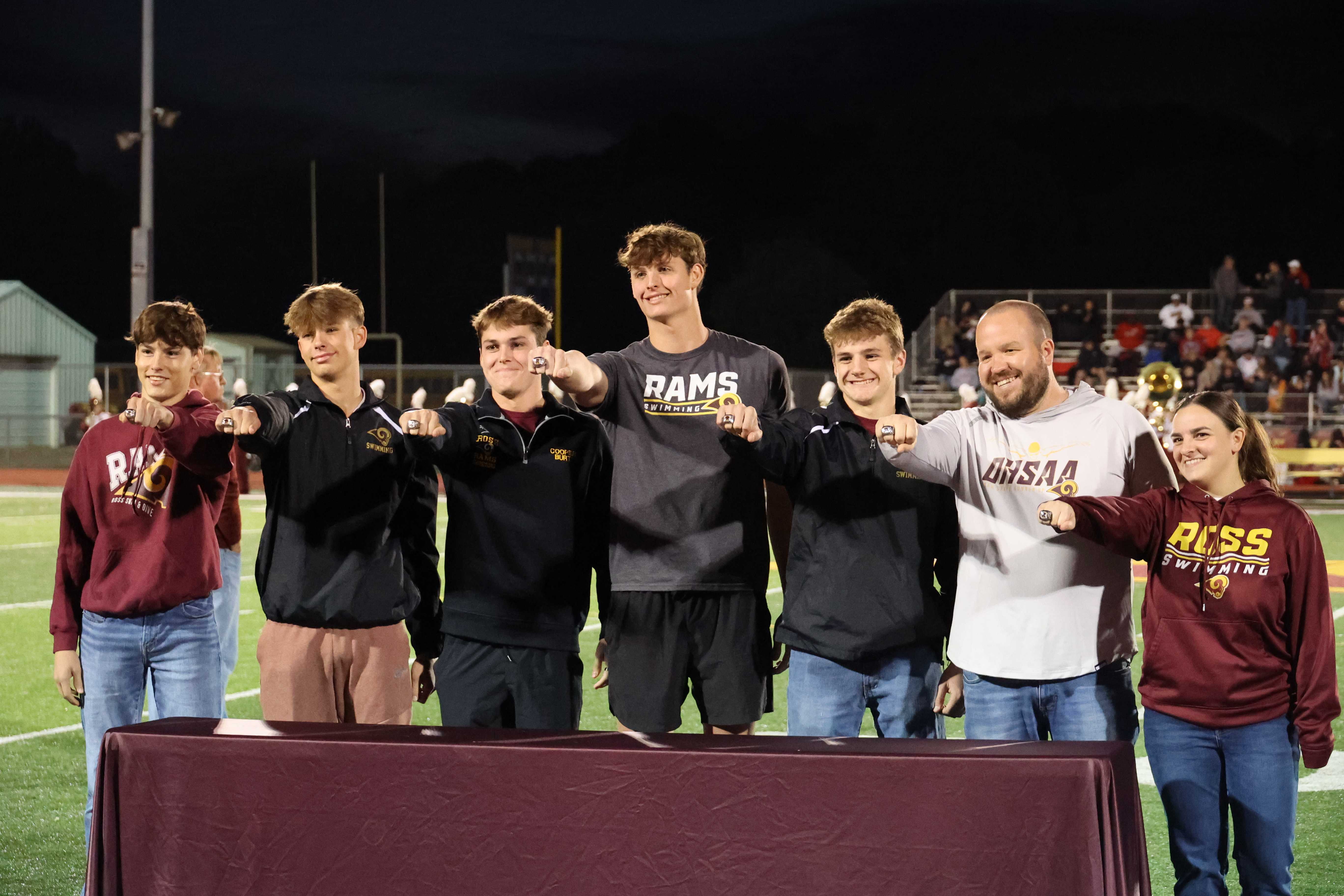 A group of young athletes and a coach stand on a football field, proudly displaying championship rings.