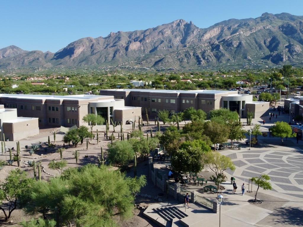 A sunny aerial view of modern low-rise buildings surrounded by trees and a paved plaza, with a dramatic mountain range in the background.