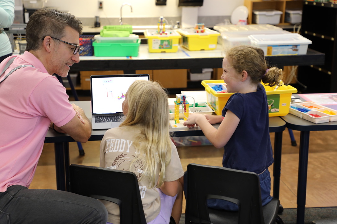 A man and two young girls work together at a table with a laptop and building blocks.