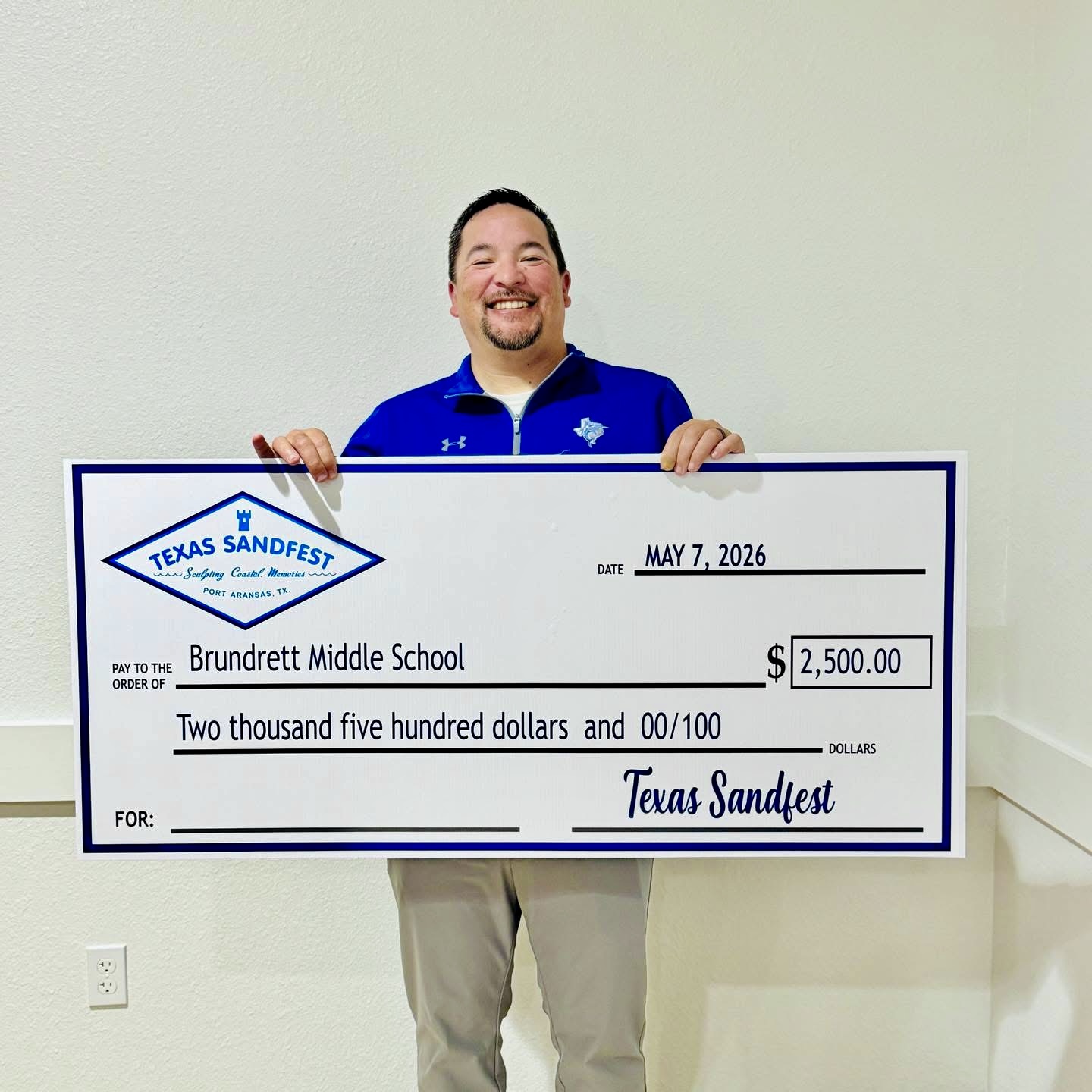 A smiling man holds a large novelty check for $2,500 made out to Broadbent Middle School.
