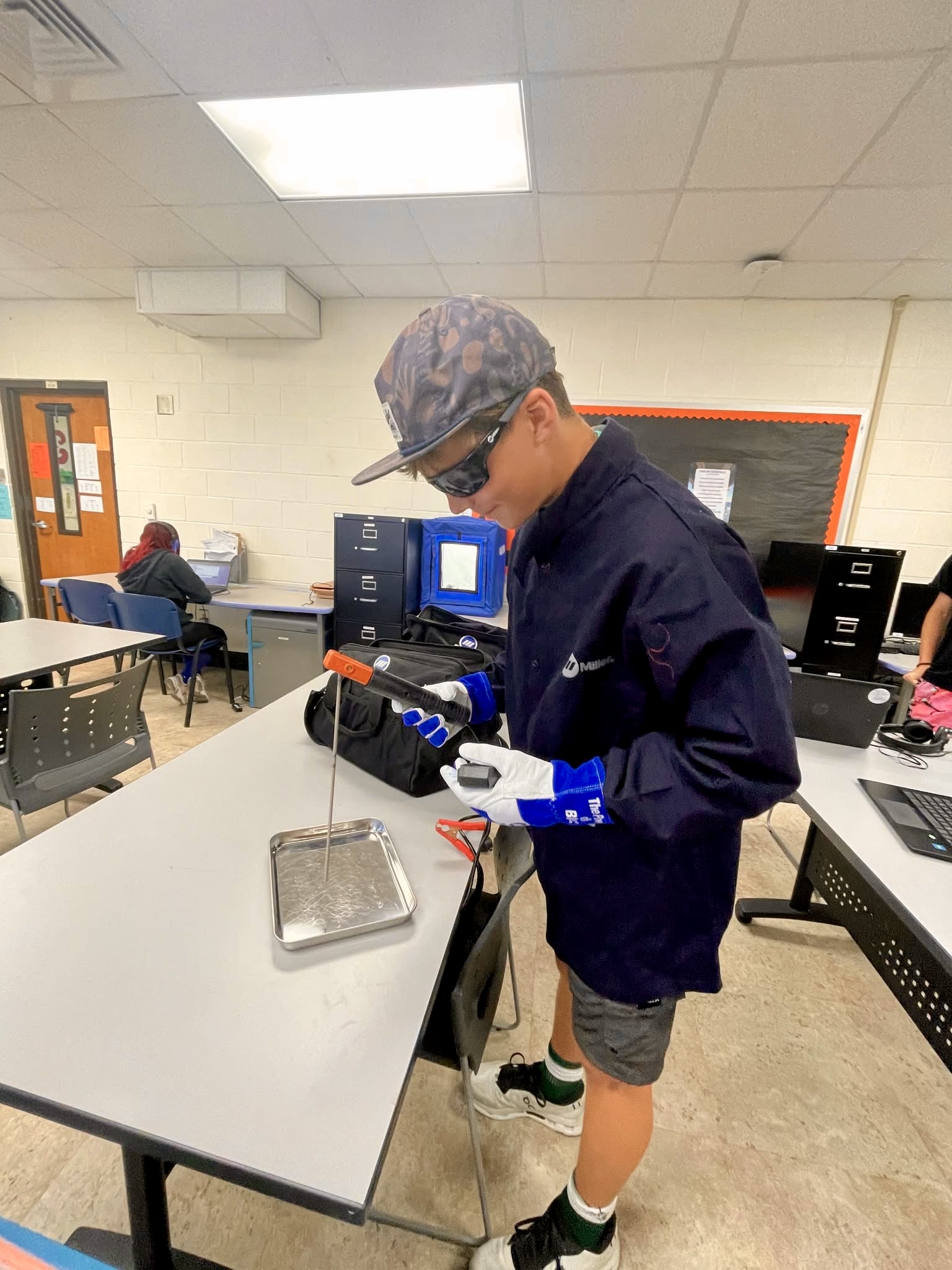 A student in a classroom wears safety glasses and gloves while working with equipment on a desk.