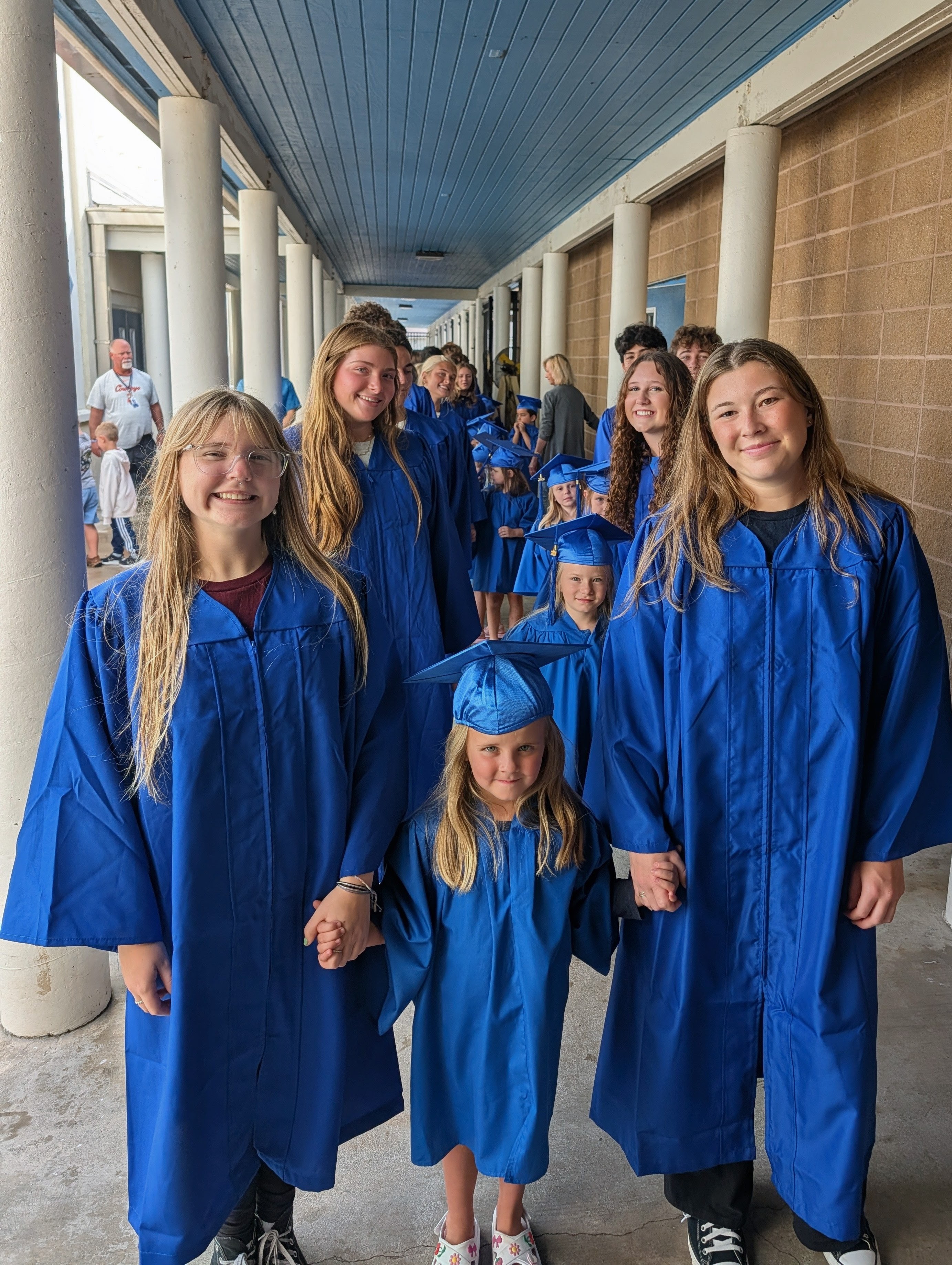 A group of smiling students in blue graduation gowns and caps walk down a hallway.