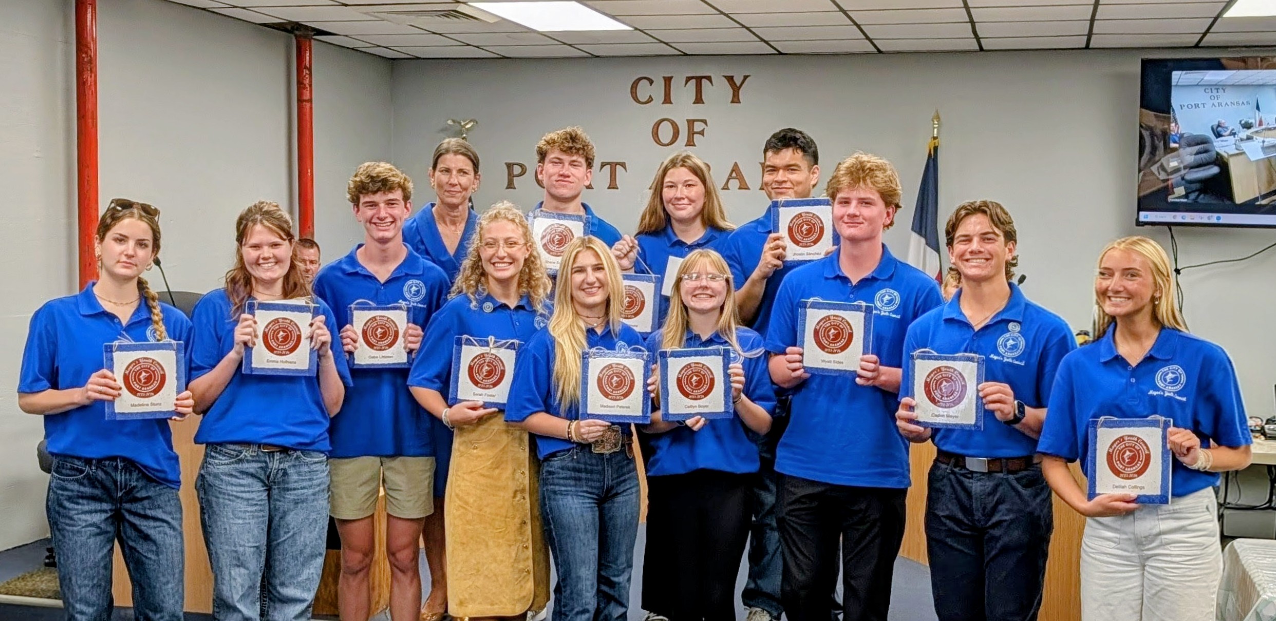 A group of young people in blue shirts stand together, holding framed certificates.
