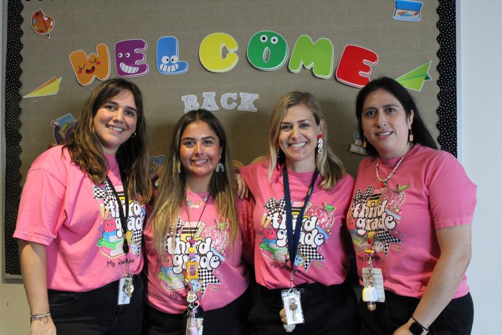 Four women in pink shirts stand in front of a bulletin board with "WELCOME BACK" text.