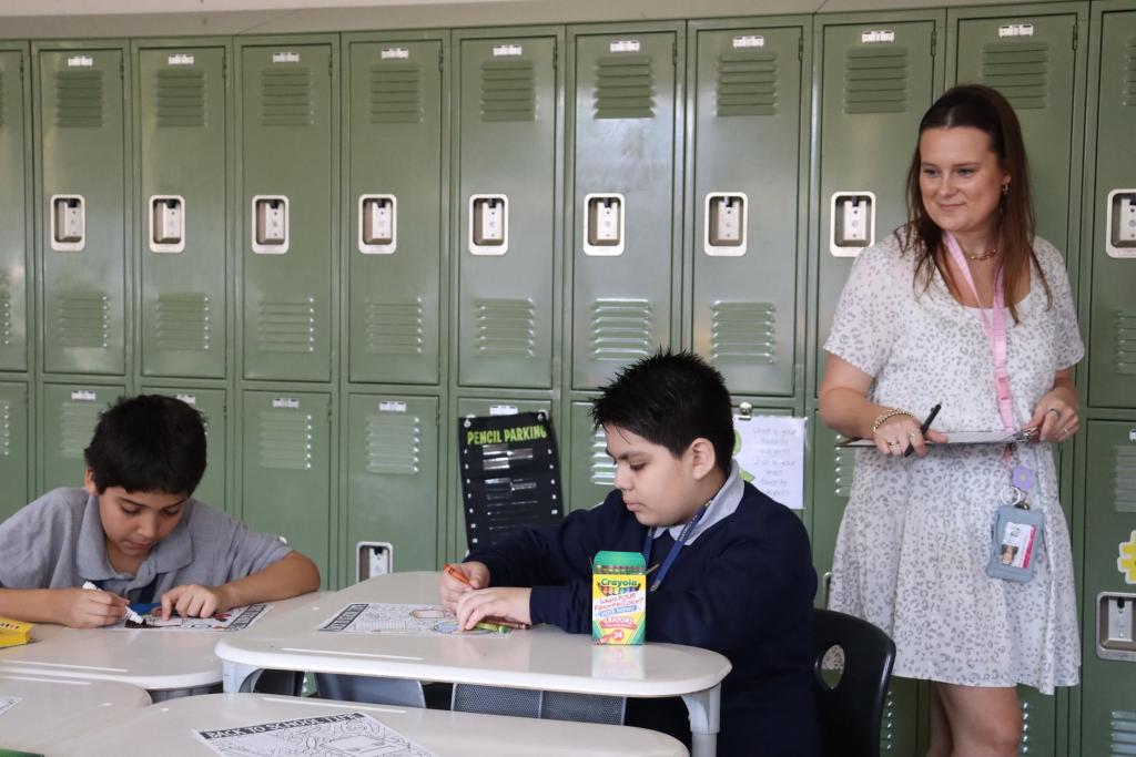 A teacher stands by two students working at desks in a classroom with lockers in the background.
