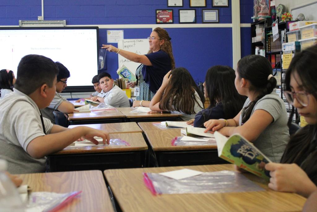 A teacher points to a projector screen while students sit at desks in a classroom.