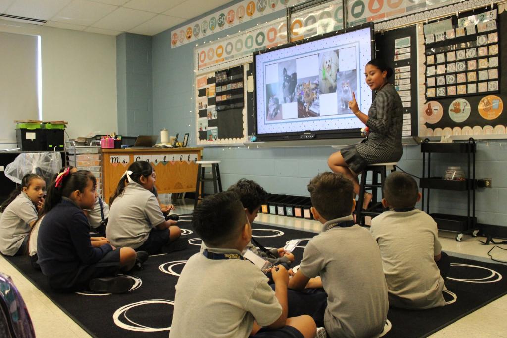 A teacher presents a lesson on a large interactive screen to a group of seated students.