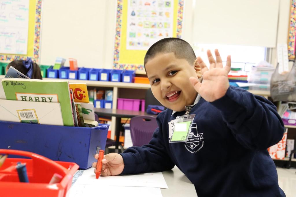A young boy waves hello from a classroom desk, holding a crayon.