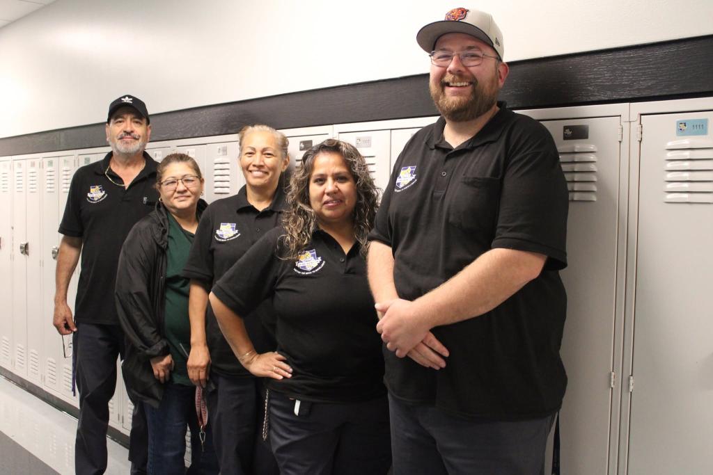 Five people in matching black shirts and hats stand in a hallway with lockers.