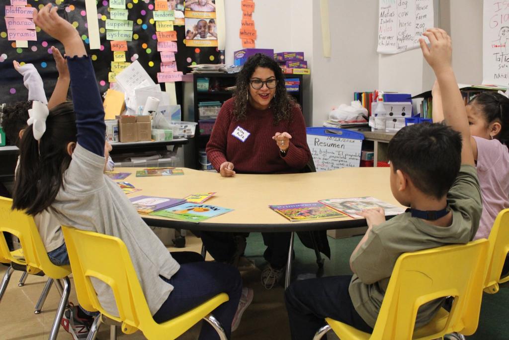 A teacher sits at a round table in a classroom, engaging with students who have their hands raised.