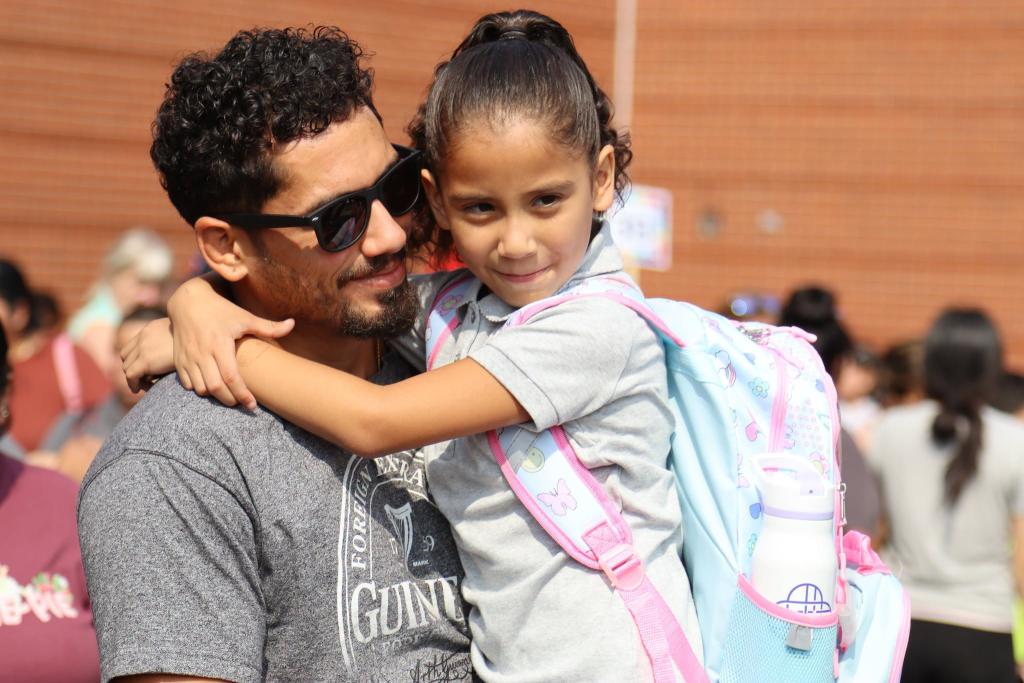A smiling young girl wearing a backpack hugs an adult outdoors in front of a brick wall.