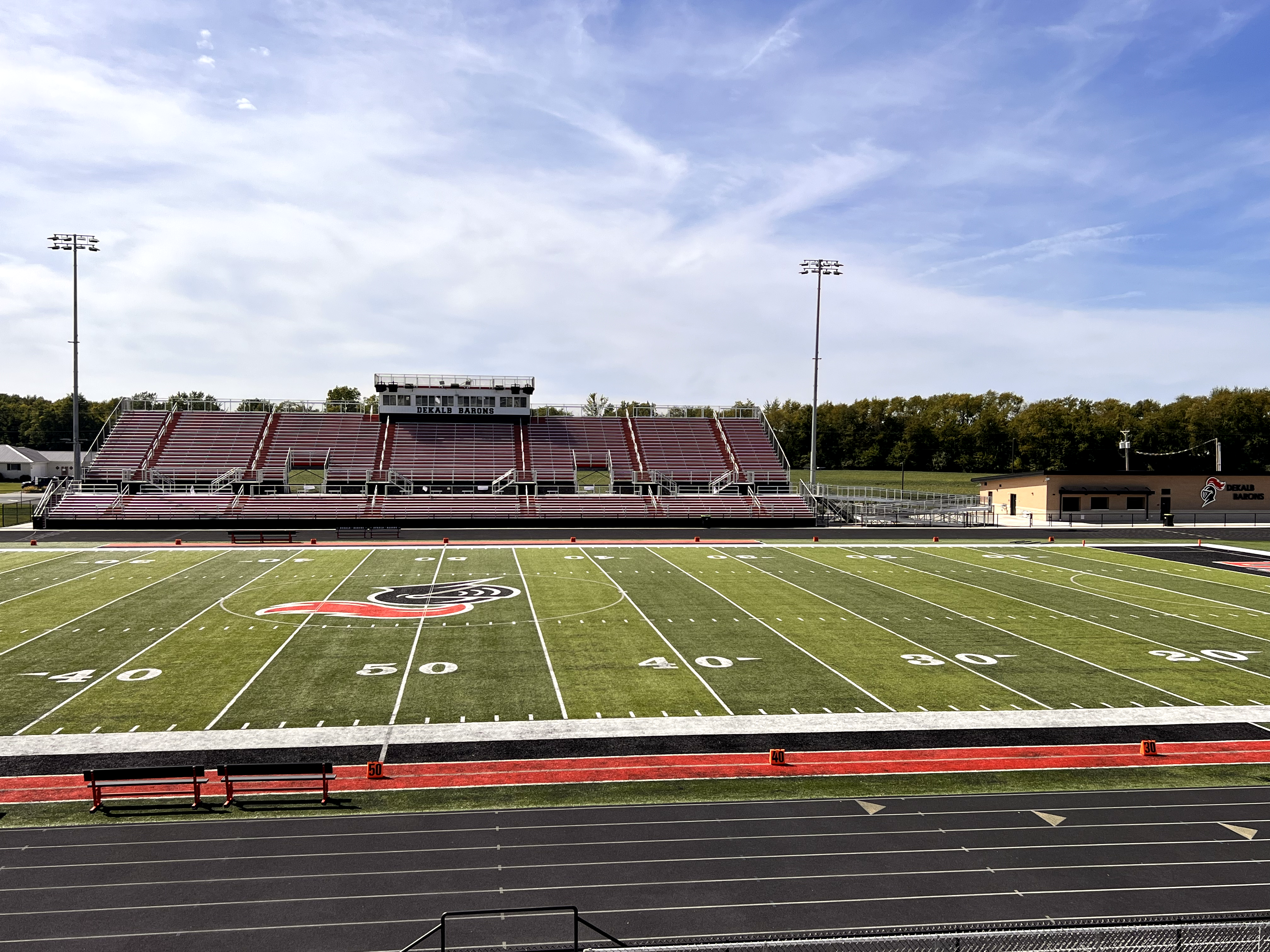 A wide-angle view of an empty american football stadium with green turf and bleachers under a blue sky.