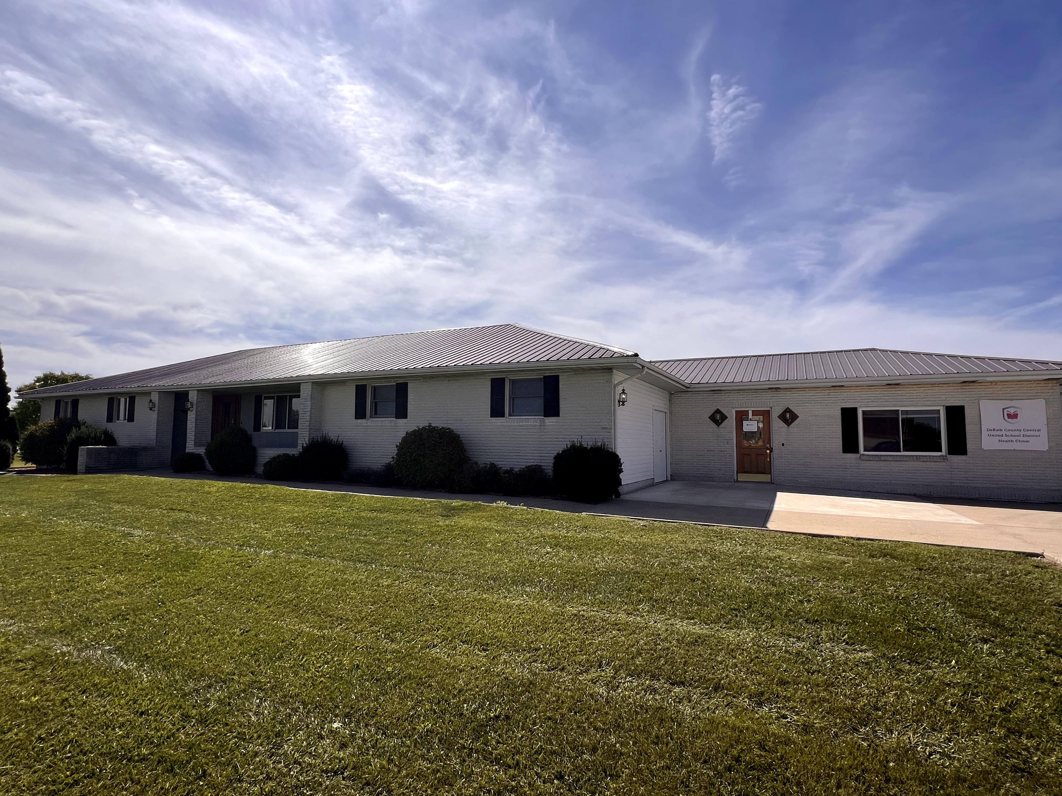 A single-story white house stands behind a well-maintained green lawn under a bright blue sky with wispy clouds.