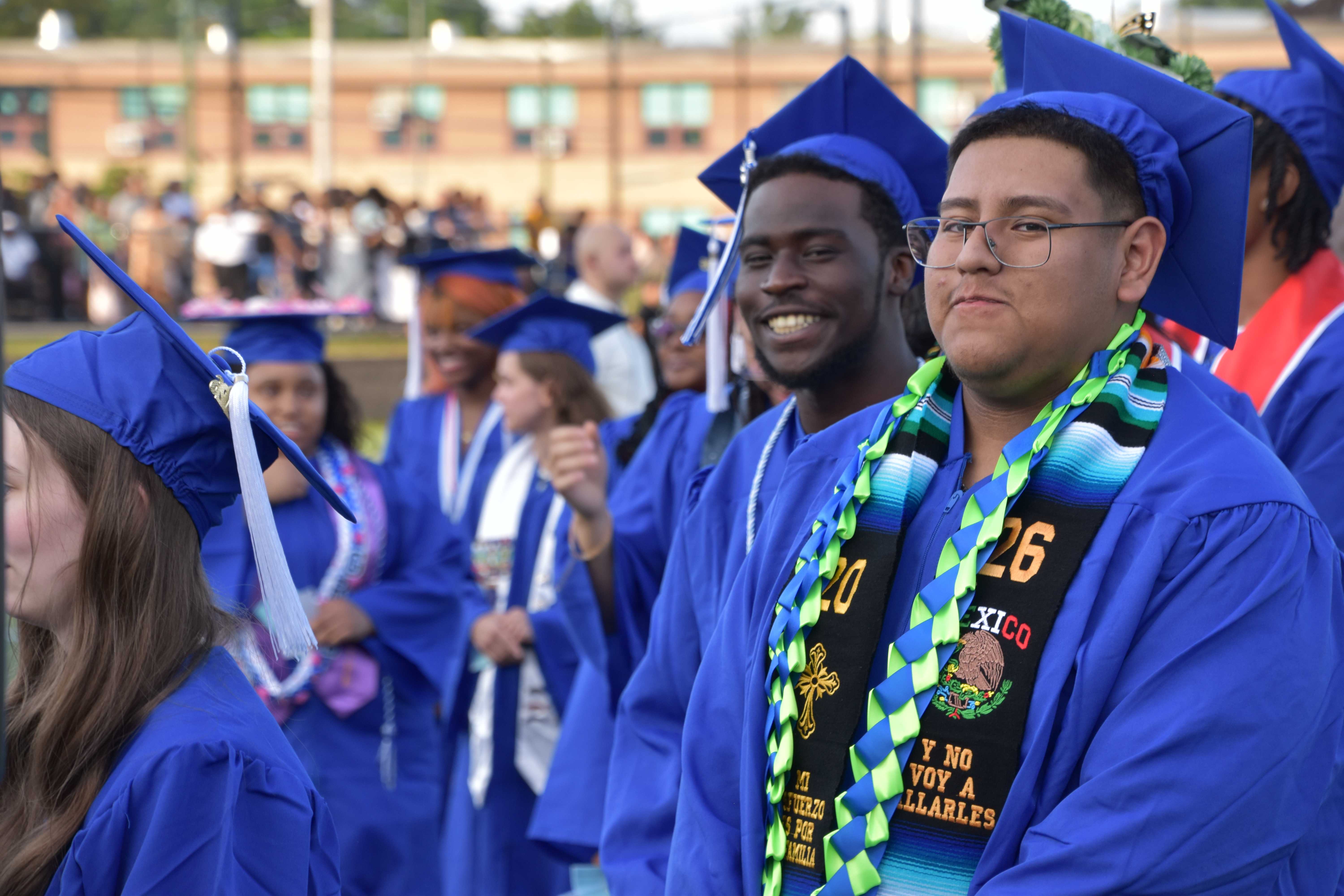 Graduates in blue caps and gowns stand in a line, some smiling, at an outdoor ceremony.