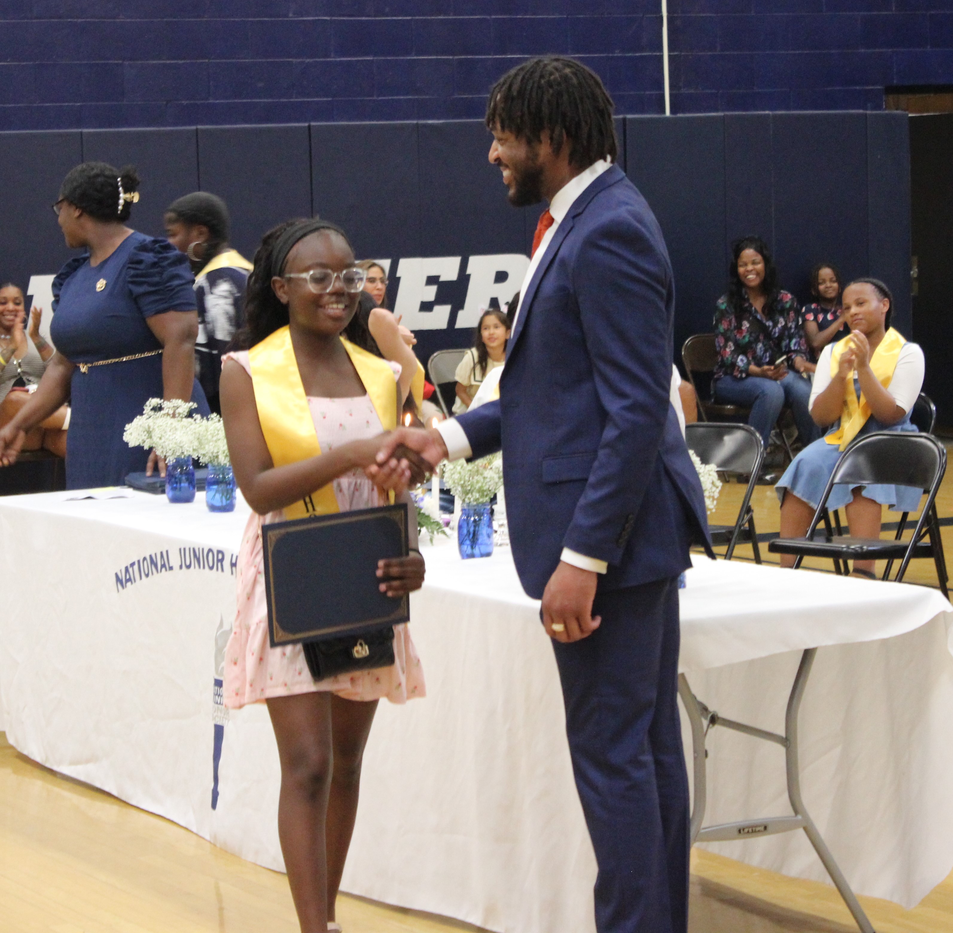 A young woman in a yellow sash shakes hands with a man in a suit at an awards ceremony.