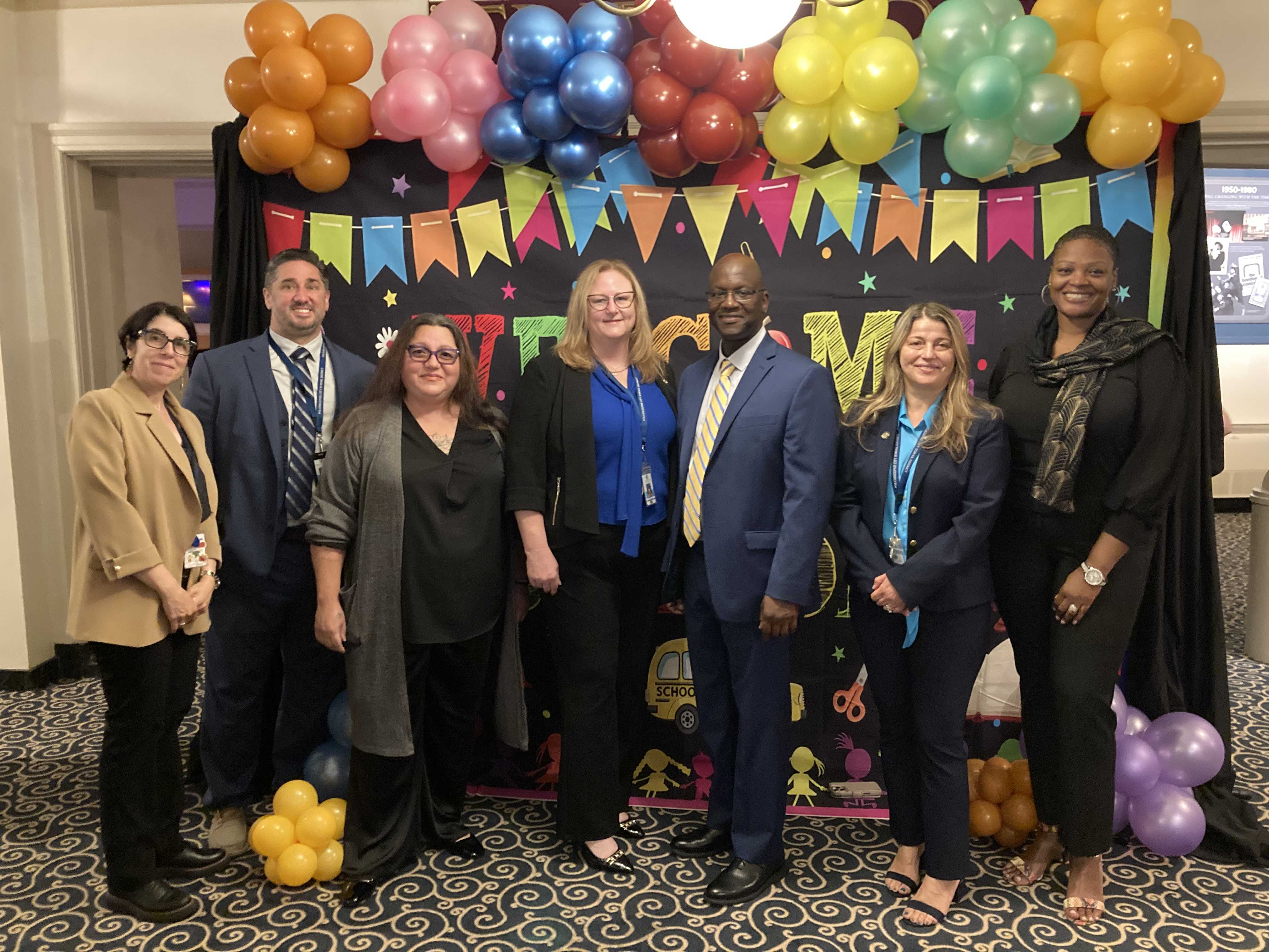 A group of people pose in front of a colorful backdrop with balloons.