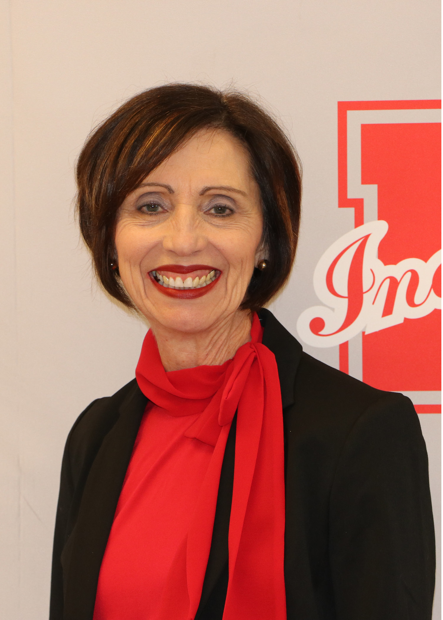 A woman with short brown hair smiles, wearing a red scarf and black blazer.