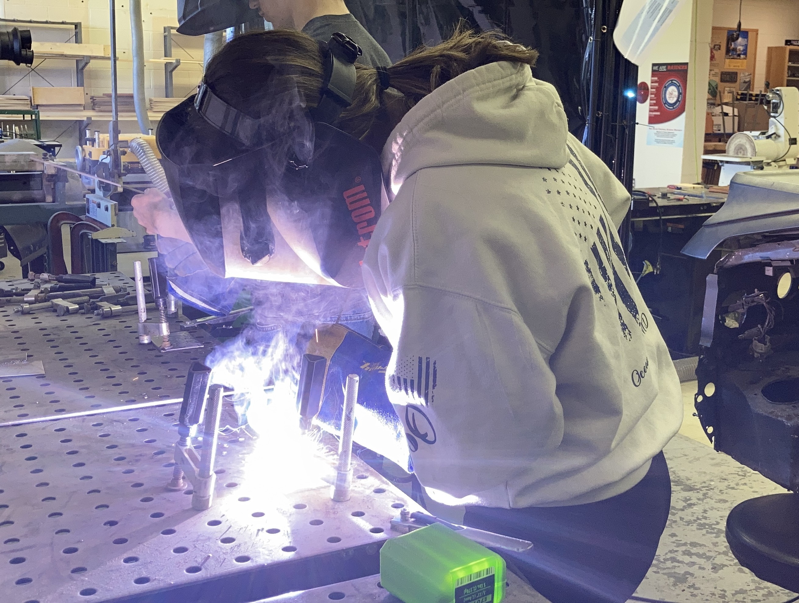 A person wearing a welding mask works on metal, creating sparks in a workshop.