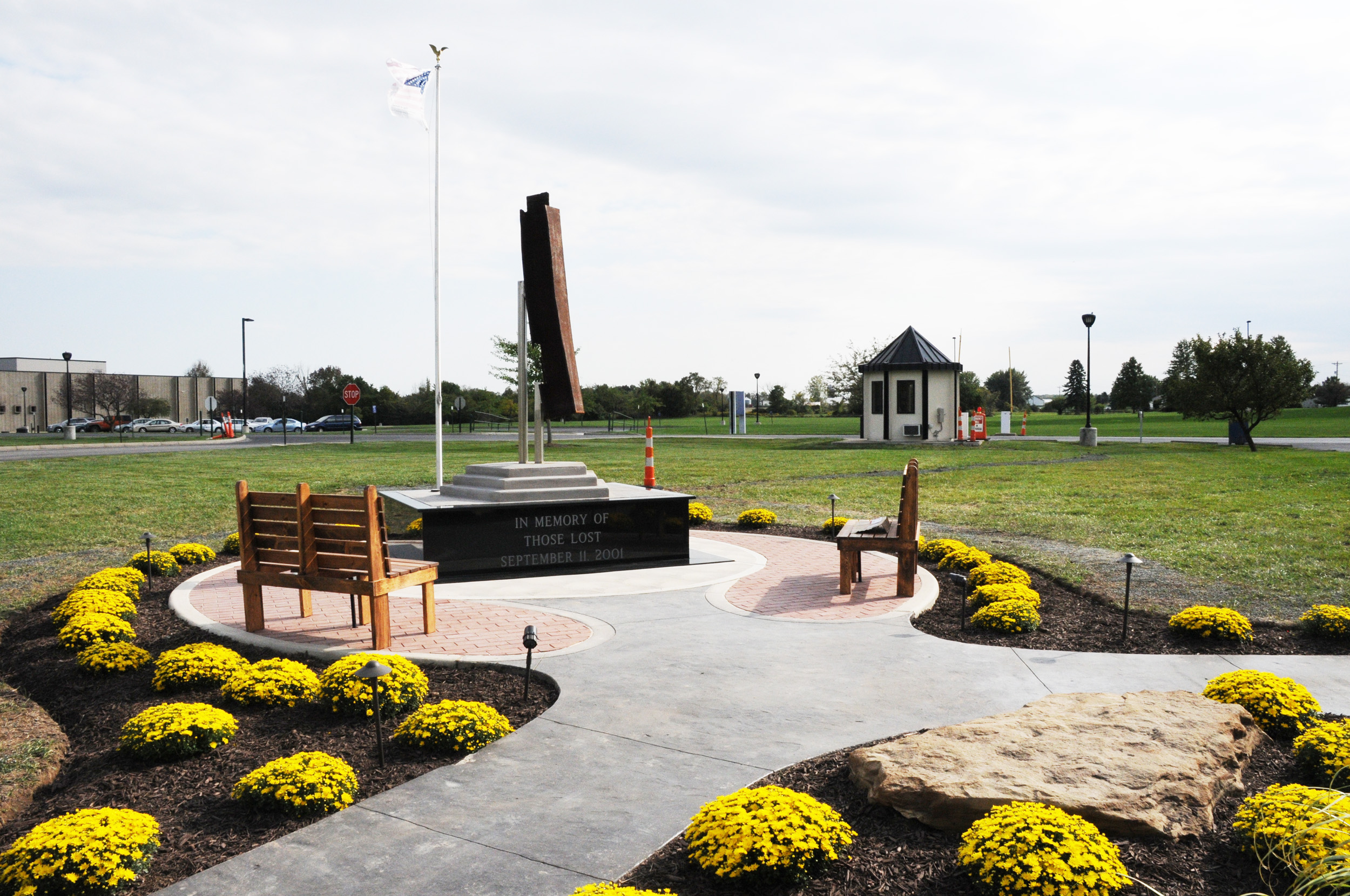A memorial with a tall, abstract sculpture and benches, surrounded by yellow flowers.