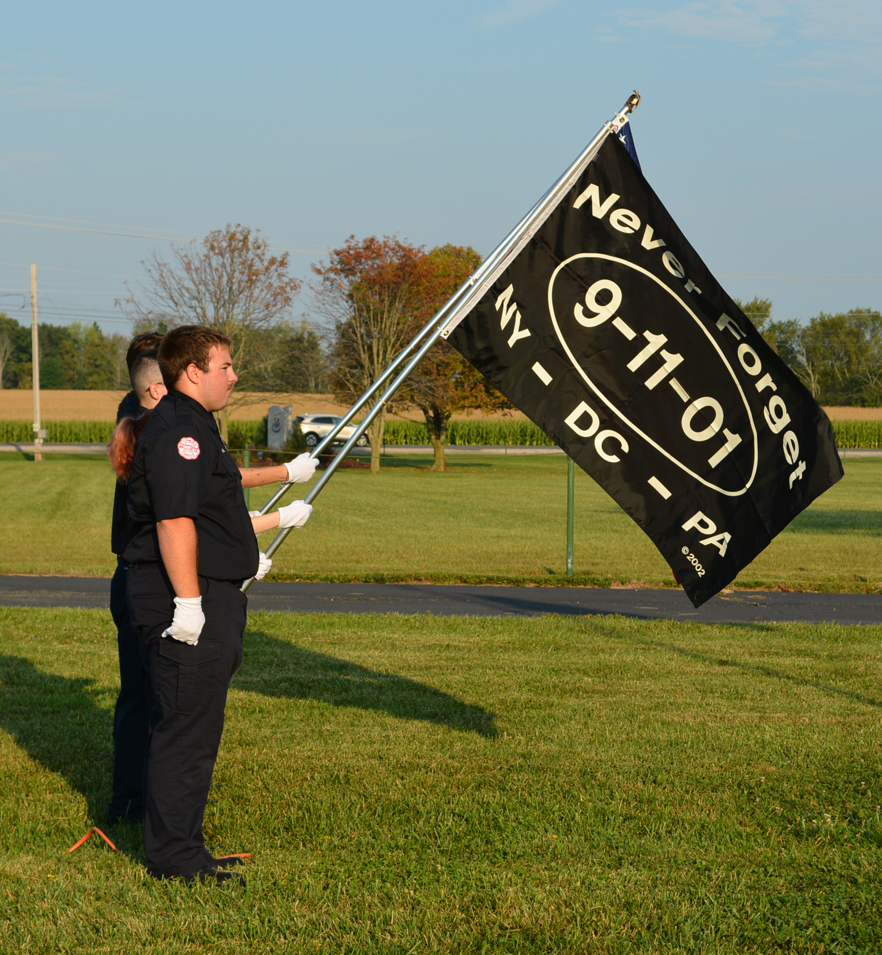 Two individuals in uniform hold a black flag with "Never Forget 9-11-01 NY - DC - PA" printed on it.