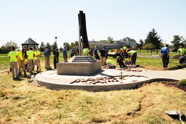 A group of people in bright yellow vests and work clothes are laying bricks around a monument.