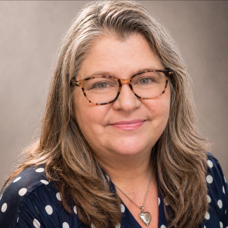 A portrait of a smiling woman with long blonde and grey hair wearing glasses and a polka dot shirt.