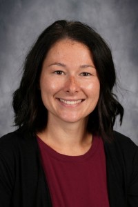 A headshot of a smiling woman with dark, shoulder-length hair and a maroon shirt against a neutral grey background.