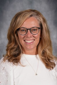 A smiling woman with blonde hair and glasses poses against a neutral gray background.