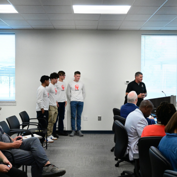 A group of young men in matching shirts stand before an audience in a conference room.