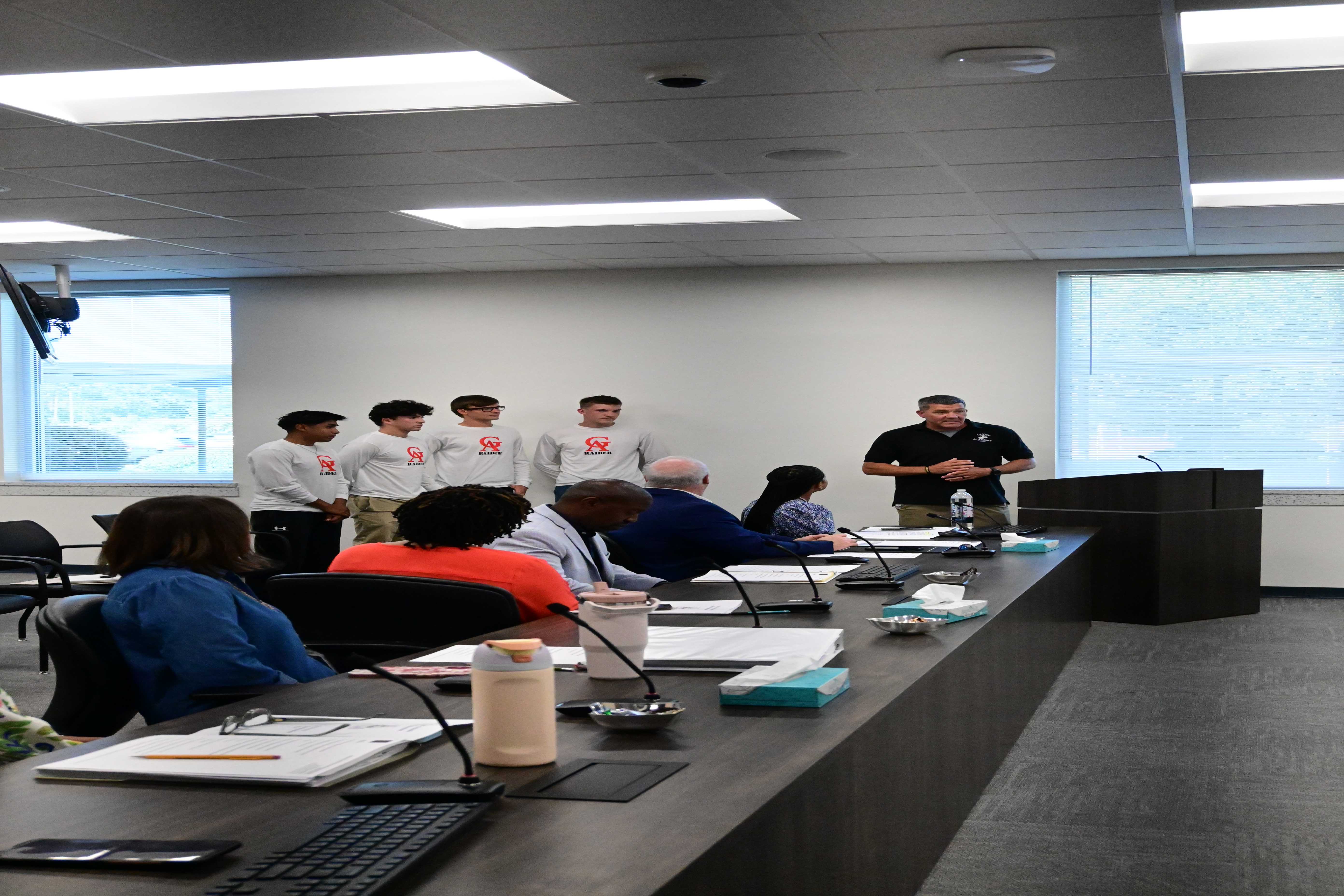 A man in a black polo shirt speaks to a group of people seated at a long table, with several young men in white shirts standing behind him.