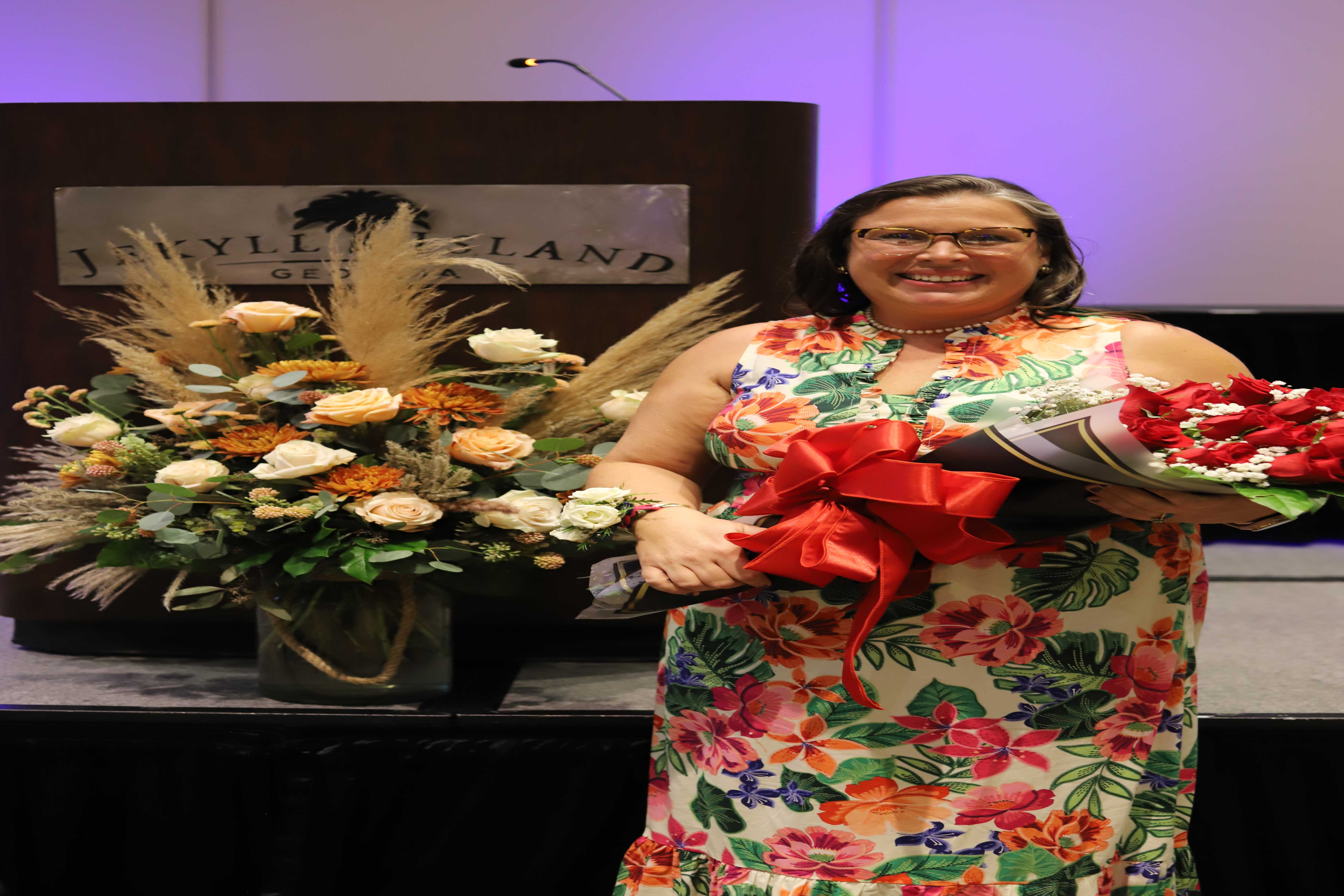 A woman in a floral dress smiles, holding bouquets of flowers at an event.