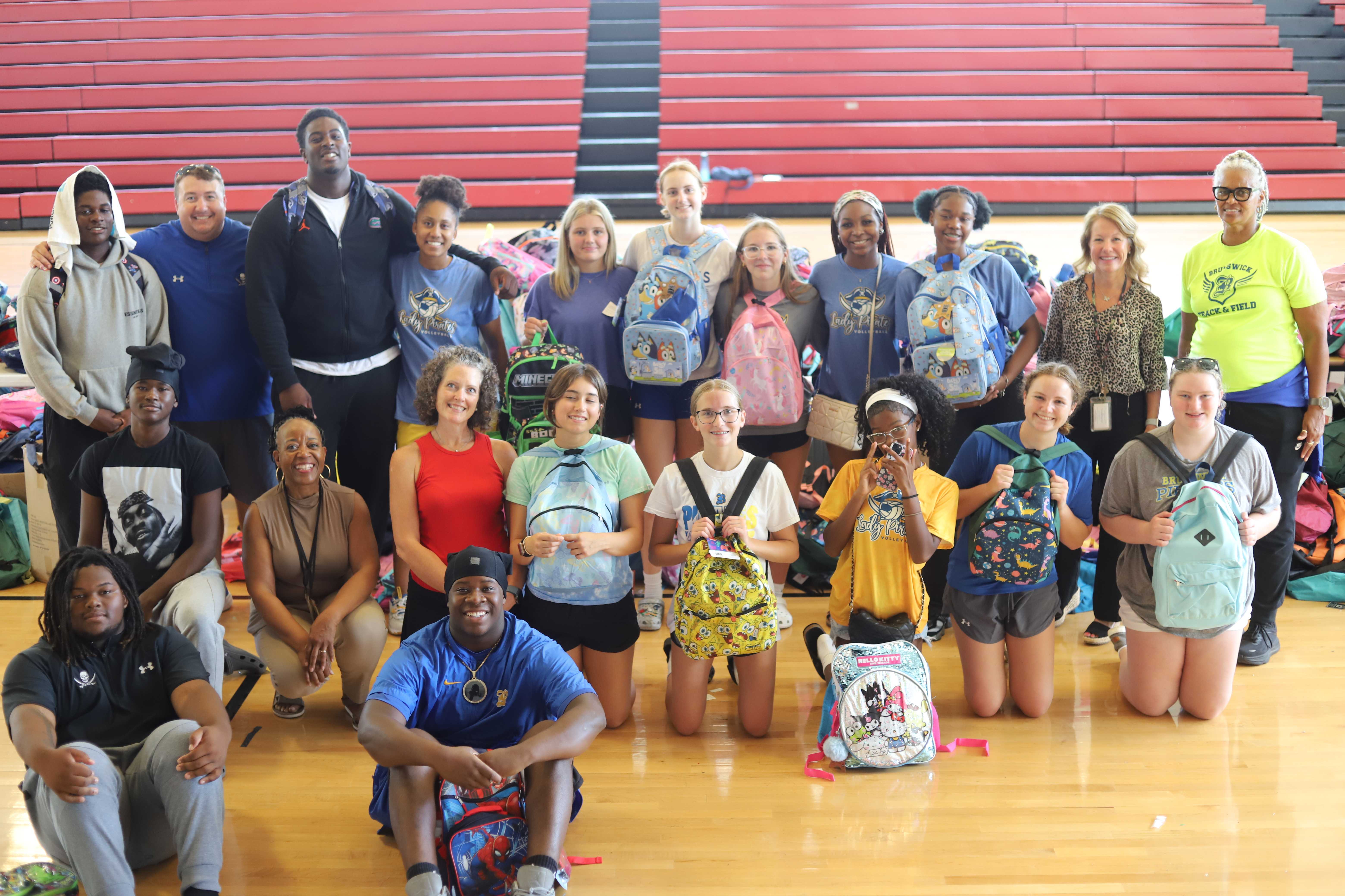 A group of student volunteers pose together, many holding backpacks, in a gymnasium.