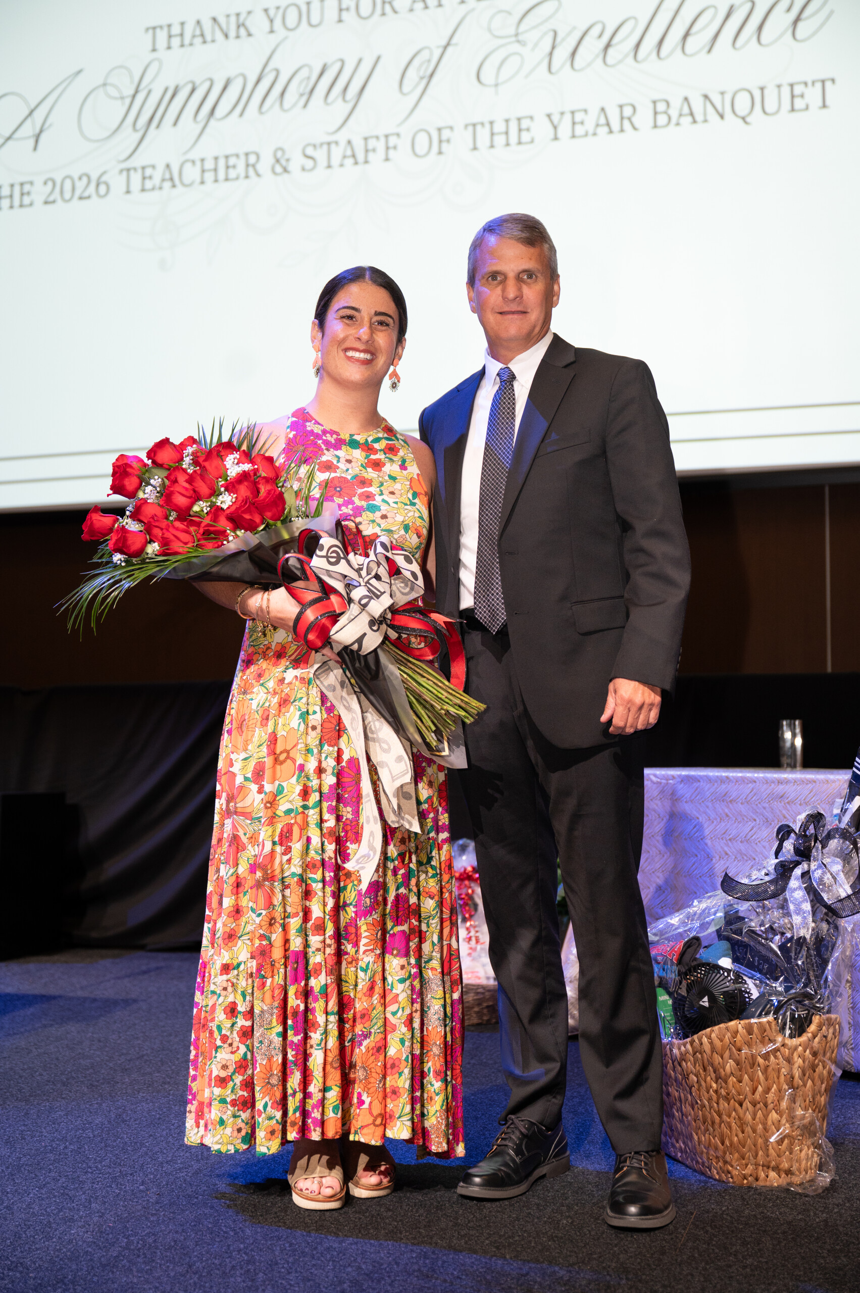 A woman in a floral dress holding red roses stands next to a man in a suit on a stage.