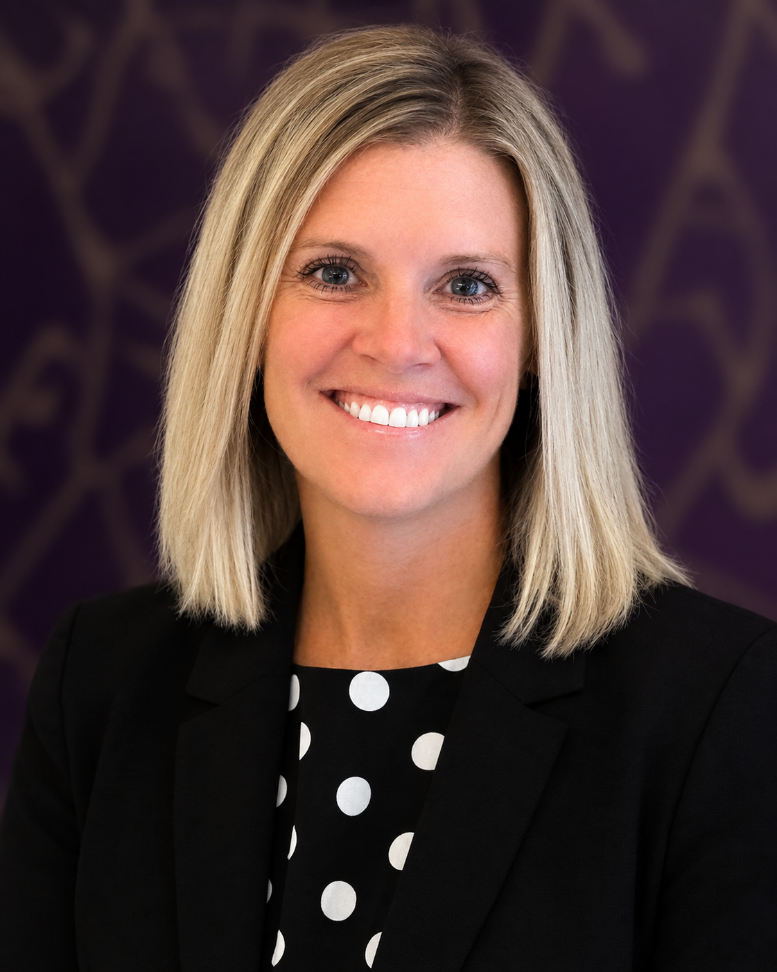 A professional headshot of a smiling woman with shoulder-length blonde hair, wearing a black blazer over a polka dot dress.