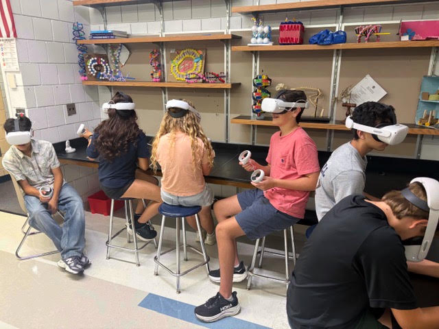 Students wearing virtual reality headsets sit on stools in a classroom with shelves.