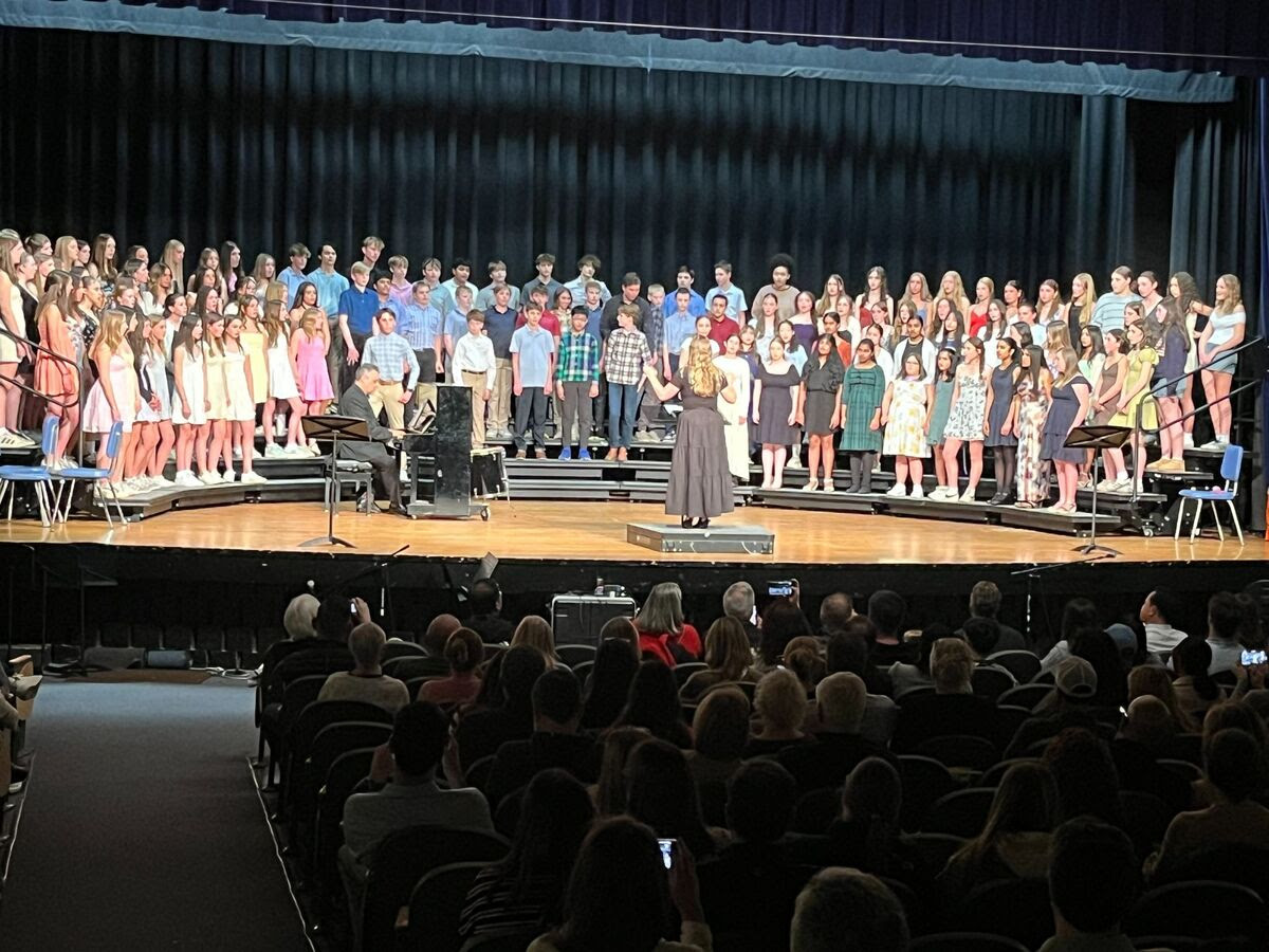 A large choir stands on a stage, performing for an audience in a dimly lit auditorium.