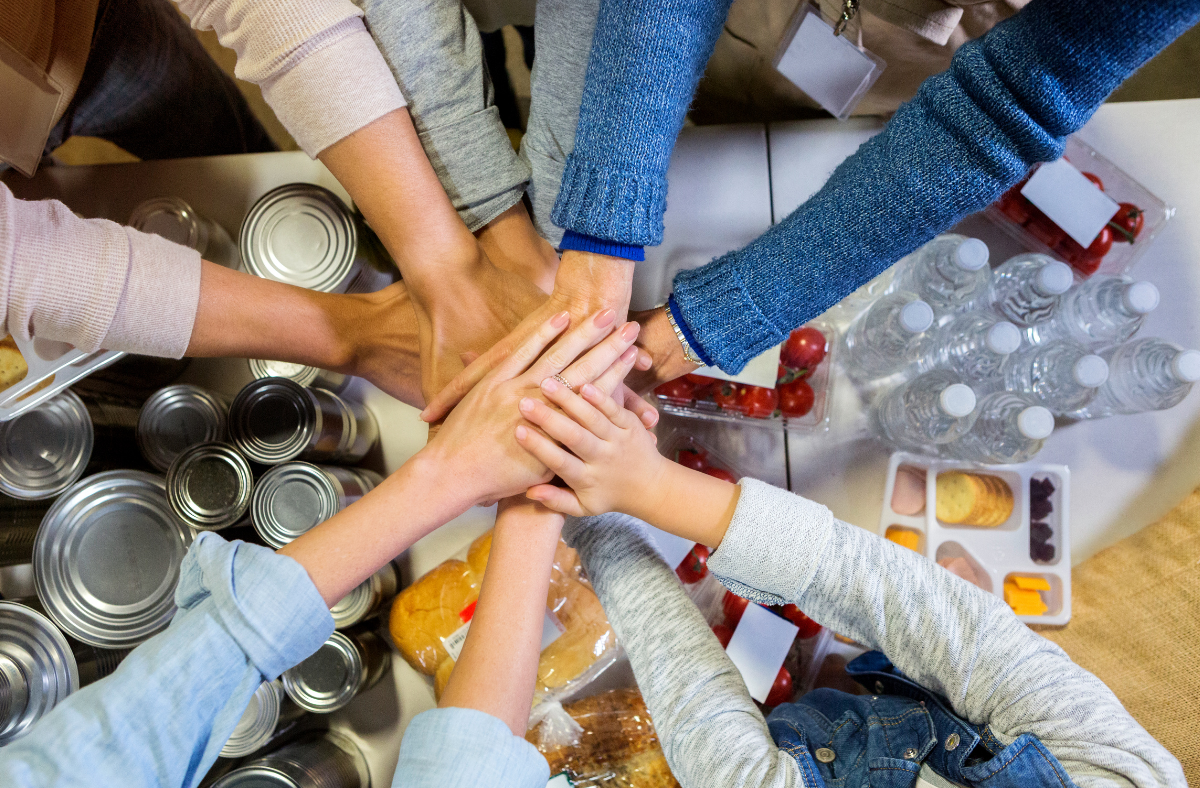 A group of hands join together in a team huddle over food donations.